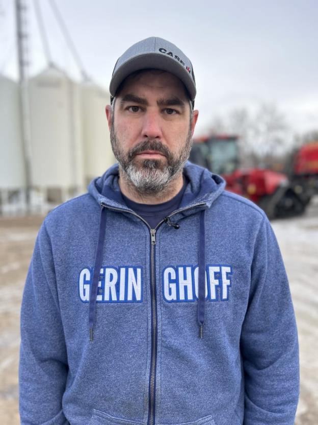 A man with a ball cap and a hooded sweater is pictured with a grain farm in the background.
